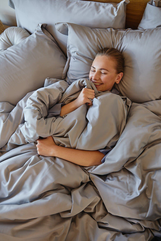 Mujer sonriente acurrucada en cama con funda edredón bamboo cotton gris-gris