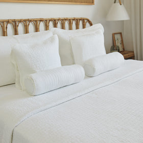 Cama con cubrecama blanco, almohadas y rollos decorativos en dormitorio-natural