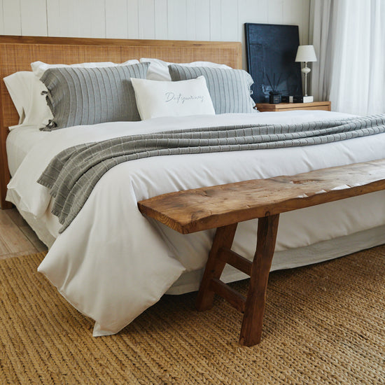 Cama con edredón blanco, almohadas grises y banco de madera al pie-blanco
