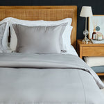 
                    
                      Cama con funda edredón gris, almohadas blancas y mesita de noche de madera-
                    
                  