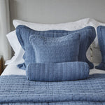 
                    
                      Almohadas azules con volantes blancos sobre cama con colcha gris-
                    
                  