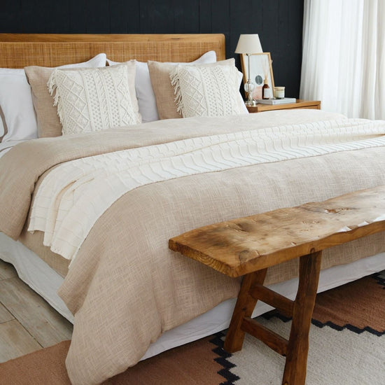 Cama con edredón beige, almohadas decorativas y banco de madera-
