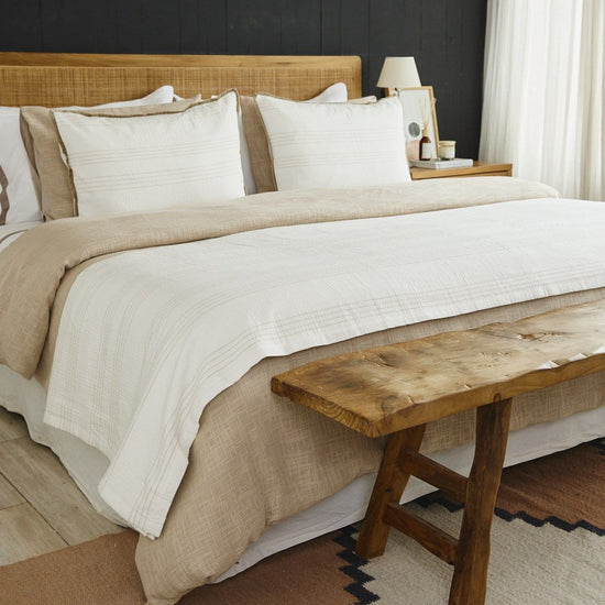 Cama con edredón blanco, almohadas y banco de madera rústica-