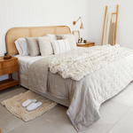 
                    
                      Cubrecama blanco sobre cama con cabecero de ratán y almohadas decorativas-
                    
                  