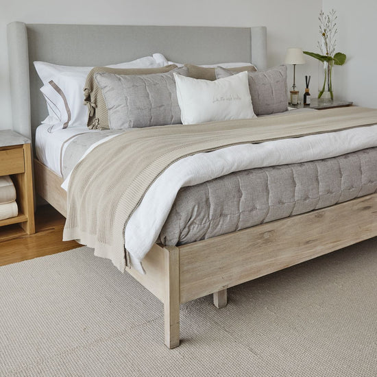 Cama con cubrecama beige, almohadas grises y cabecero tapizado gris-beige