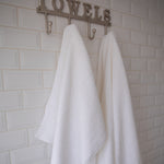 
                    
                      Dos toallas blancas colgadas en ganchos bajo letrero "TOWELS-blanco
                    
                  