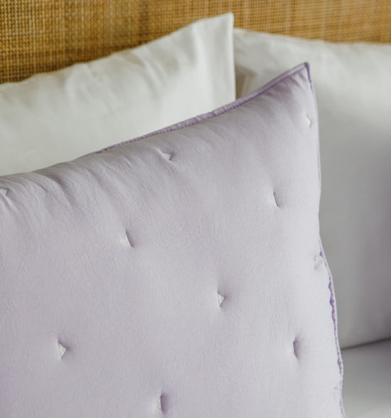 Almohada lila con botones decorativos sobre cama blanca-lila