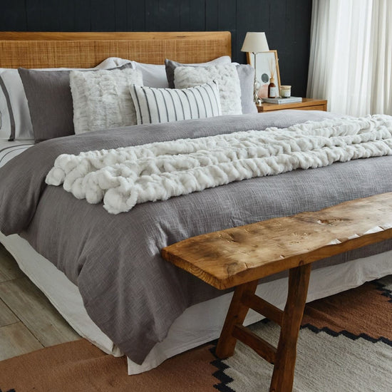 Cama con edredón gris, almohadas decorativas y banco de madera-