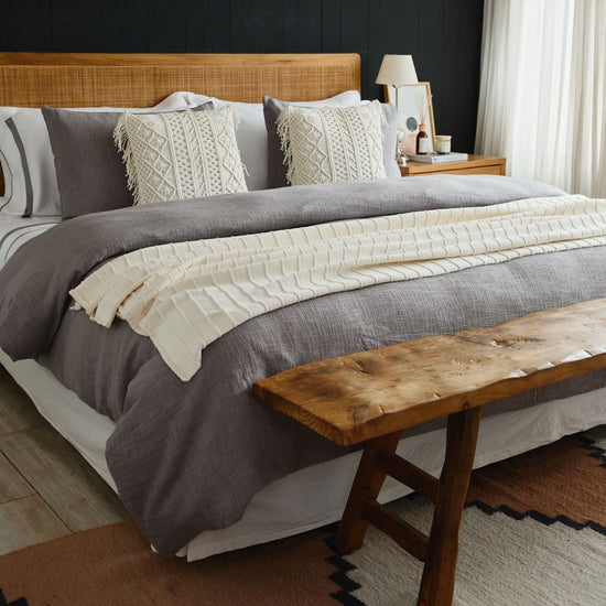 Cama con funda de edredón gris, almohadas decorativas blancas y banco de madera-