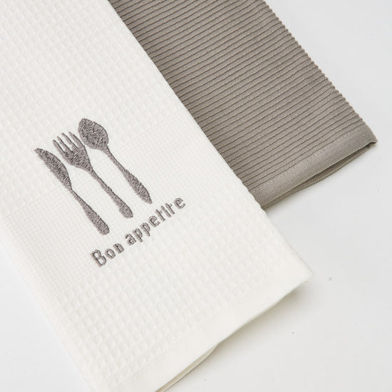 Dos repasadores, uno blanco con cubiertos y texto Bon appetit, otro gris-gris
