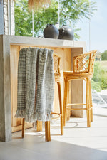 
                    
                      Manta Santorini gris colgada en mostrador de madera con sillas de ratán-gris
                    
                  