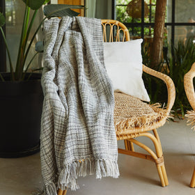 Manta gris Santorini draped sobre silla de ratán con cojín blanco-gris