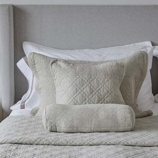 Almohada y cilindro decorativo beige sobre cama con cabecera gris-beige