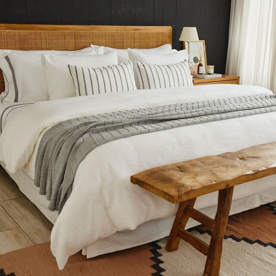Cama con edredón gris, almohadas blancas y banco de madera-