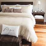 
                    
                      Cama elegante con edredón beige, almohadas decorativas y mesita de noche de madera-
                    
                  