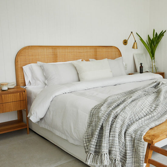 Manta Santorini gris sobre cama con cabecero de ratán y ropa blanca-gris